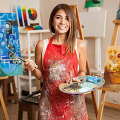 A smiling person stands in an art studio, wearing a paint-splattered red apron, holding brushes and a palette, surrounded by colorful abstract paintings