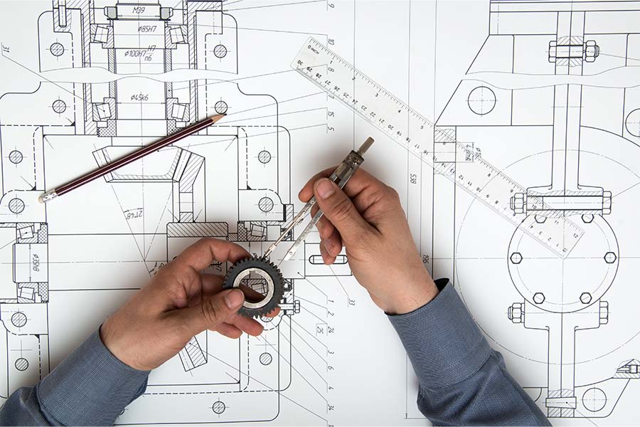 Hands rest on blueprints holding a gear and scaling tool