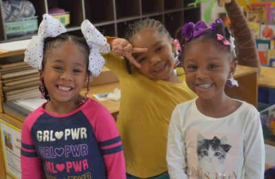 Three children with blurred faces in a classroom; one wears a 'GRL PWR' shirt, another a yellow shirt making a peace sign, and the third a cat-themed shirt.