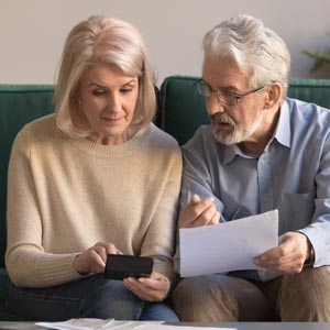 Retired couple sitting on a couch, one holding a smartphone and the other holding a piece of paper.