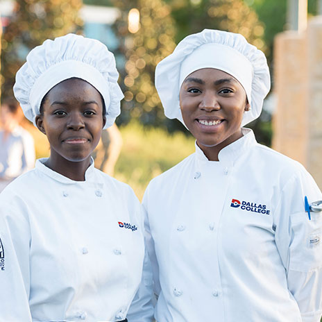 Two culinary students wearing white hats and chef uniforms with the Dallas College logo, standing side by side outdoors.