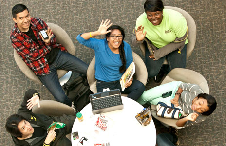 Overhead view of five students sitting around a circular table with a laptop, snacks, and drinks, engaged in reading, using phones, and interacting.
