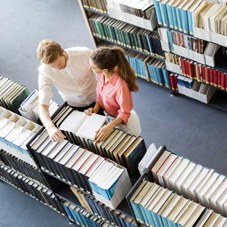 Two people in a library looking at an open book together, surrounded by bookshelves, viewed from above