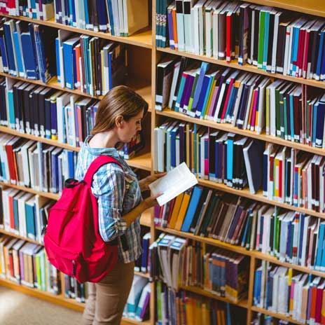 Student with red backpack, reading a book in front of a library bookshelf