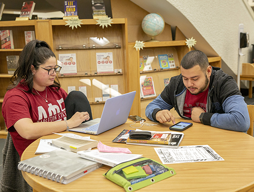 Two students working at a round table in a study room, surrounded by a laptop, notebooks, papers, and stationery.