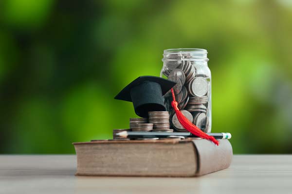 Jar filled with coins, a small graduation cap on a stack of coins, and an old book, set against a green outdoor background.