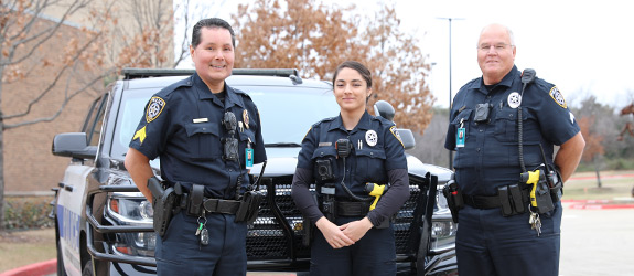 Three campus police officers in dark blue uniforms standing in front of a police vehicle, with trees and a building in the background.