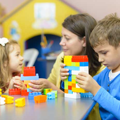 A teacher sits at a desk with two students and plays with large LEGO blocks