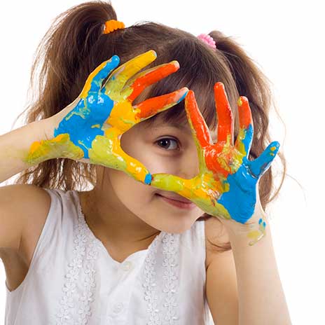 A young girl holds her hands in front of face with her fingers spread out and a variety of paint colors covering her hands
