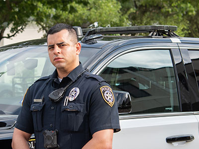 A police officer in uniform stands confidently in front of a patrol car