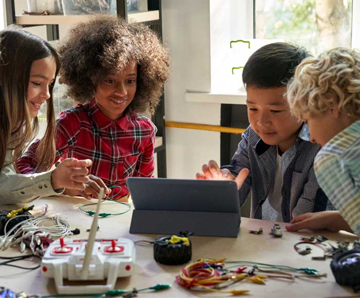 Four children sitting at a table engaged with a tablet, surrounded by electronic components and wires, smiling and focused, conveying collaboration and curiosity