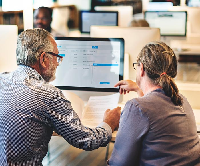 A man and woman sit in front of a computer, discussing a document in a bright office