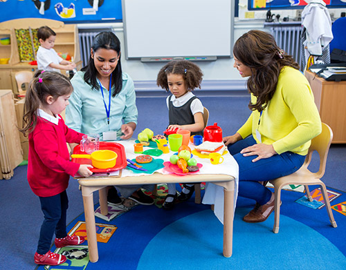 Two teachers sit at a small table with pre-school students while they play with toys